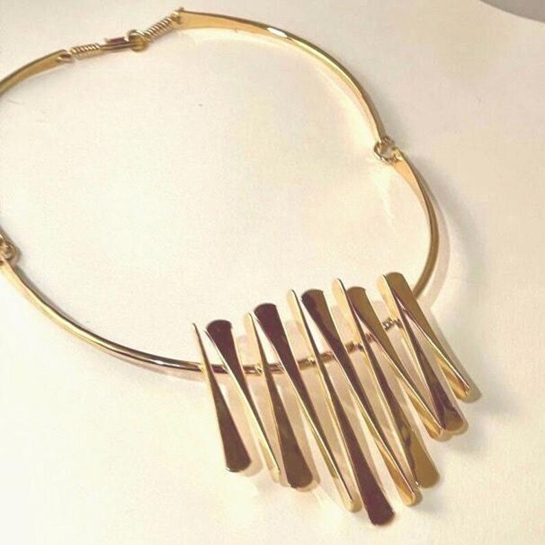 Pode incluir: Colar dourado com gola r&iacute;gida e curva e um pendente composto por barras verticais e alongadas. As barras est&atilde;o dispostas em um padr&atilde;o escalonado, criando um design geom&eacute;trico. O colar tem um fecho.