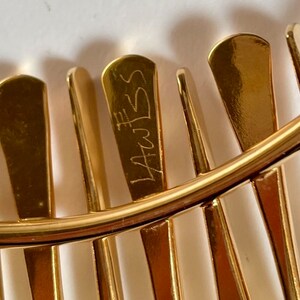 Pode incluir: Close-up de uma joia de metal dourado com um design curvo. A pe&ccedil;a apresenta elementos retangulares verticais e uma faixa curva. A inscri&ccedil;&atilde;o "LAWSS" &eacute; vis&iacute;vel em um dos elementos retangulares. A joia tem um acabamento polido.