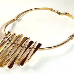 Pode incluir: Um colar dourado com uma gola r&iacute;gida e curva e um grupo de barras planas, verticais e alongadas. As barras s&atilde;o dispostas em um padr&atilde;o escalonado, criando um efeito em camadas. O colar tem um fecho simples.