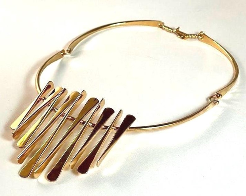 Pode incluir: Um colar dourado com uma gola r&iacute;gida e curva e um grupo de barras planas, verticais e alongadas. As barras s&atilde;o dispostas em um padr&atilde;o escalonado, criando um efeito em camadas. O colar tem um fecho simples.