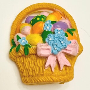 May include: A yellow basket-shaped decoration filled with colorful Easter eggs. The eggs are various colors, including green, orange, yellow, and purple. A light blue flower arrangement and a pink bow adorn the front of the basket. The basket has a woven texture.