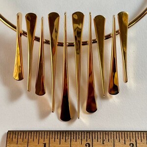 Pode incluir: Um colar dourado com uma s&eacute;rie de pingentes verticais, alongados, em forma de gota. Os pingentes variam em tamanho e s&atilde;o presos a uma fina corrente dourada curva. O colar &eacute; exibido em uma superf&iacute;cie branca com uma r&eacute;gua de madeira.