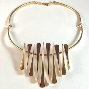 Pode incluir: Um colar dourado com uma gola r&iacute;gida e curva e uma s&eacute;rie de pingentes verticais, alongados e em forma de gota. Os pingentes s&atilde;o dispostos em uma fileira, suspensos de uma barra horizontal. O colar tem um fecho simples.
