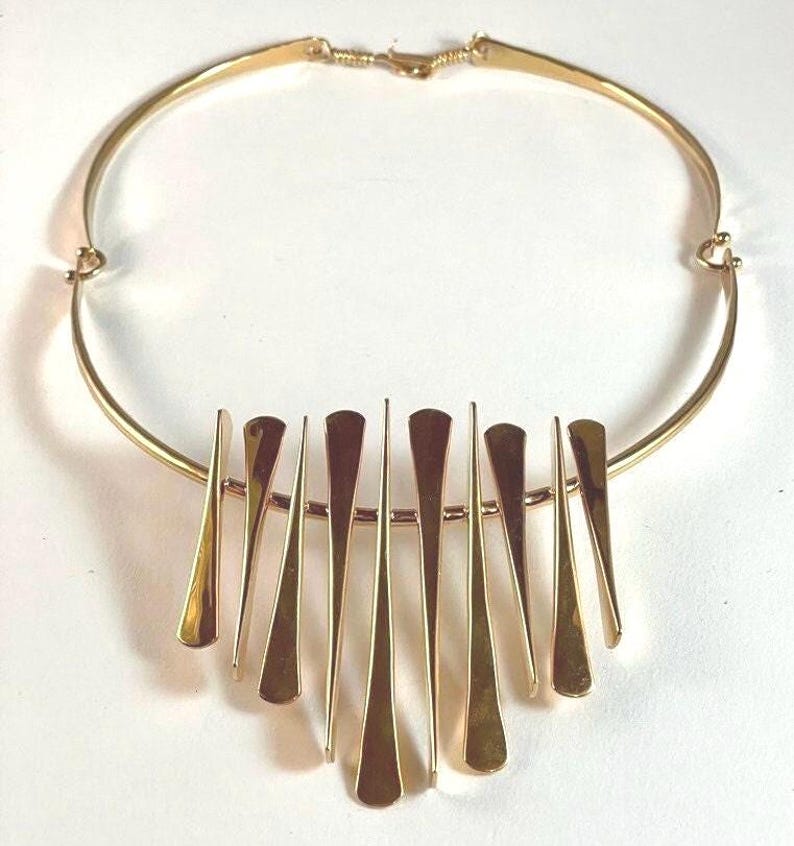 Pode incluir: Um colar dourado com uma gola r&iacute;gida e curva e uma s&eacute;rie de pingentes verticais, alongados e em forma de gota. Os pingentes s&atilde;o dispostos em uma fileira, suspensos de uma barra horizontal. O colar tem um fecho simples.