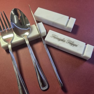 May include: A fork, spoon, and knife rest on white marble holders. The spoon and fork are silver with beaded edges. One holder has the text "Famiglia Filippi". The items are on a maroon surface.