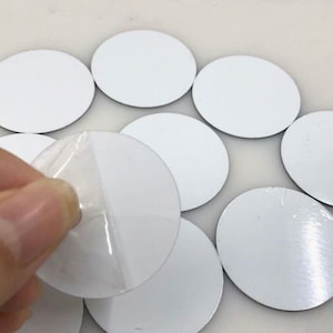 May include: A collection of white, oval-shaped stickers with a peel-off backing. One sticker is being peeled, revealing the adhesive. The stickers have a smooth, reflective surface and are arranged on a light-colored background.