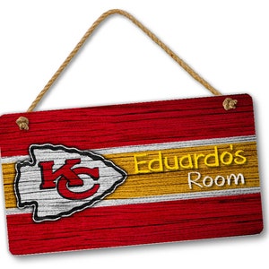 May include: A wooden sign with a distressed woodgrain finish. The sign features the Kansas City Chiefs logo in red and white with a yellow and white stripe. The text "Eduardo's Room" is written in yellow letters on the sign.