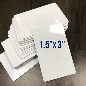May include: A stack of rectangular white plastic cards with rounded corners. The top card features the blue printed text 1.5" x 3", indicating the dimensions of the cards. The cards have a smooth, glossy finish and are arranged in a slightly fanned pile.