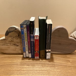 May include: A pair of heart-shaped wooden bookends, one light brown and one gray, holding up a collection of books and DVDs. The books include titles like "Hearing God", "Boo to You", and "The Final Lectures". The DVDs include titles like "Mary Poppins", "Cinderella", and "Madeline".