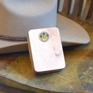 May include: A light-coloured wooden hat rack with a metal keyhole plate for hanging. The rack is rectangular and has a natural wood grain. A tan cowboy hat is in the background, resting on a wooden chair.