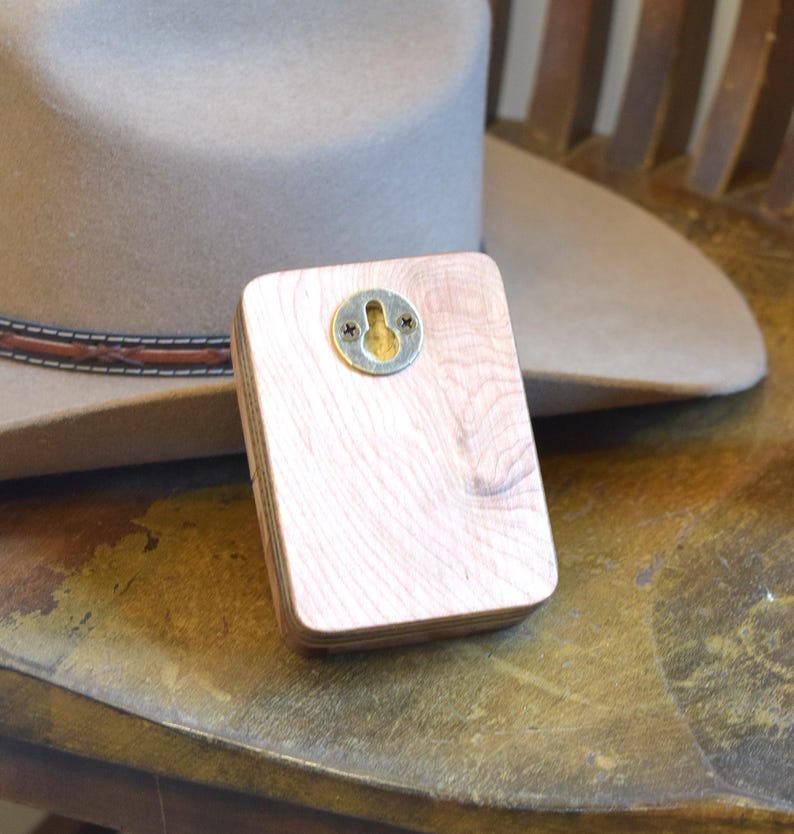 May include: A light-coloured wooden hat rack with a metal keyhole plate for hanging. The rack is rectangular and has a natural wood grain. A tan cowboy hat is in the background, resting on a wooden chair.