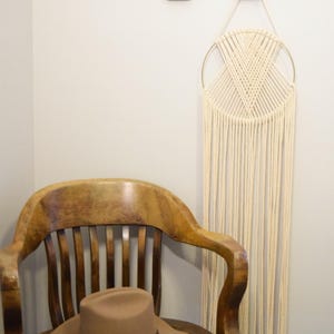 May include: A tan cowboy hat sits on a wooden chair. A cream-coloured macrame wall hanging with a gold ring hangs on the wall. Two decorative turquoise and brown wall hooks are above the macrame.