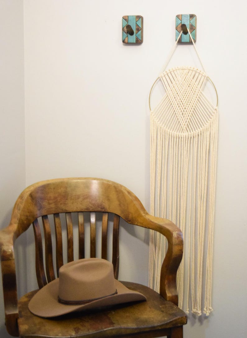 May include: A tan cowboy hat sits on a wooden chair. A cream-coloured macrame wall hanging with a gold ring hangs on the wall. Two decorative turquoise and brown wall hooks are above the macrame.