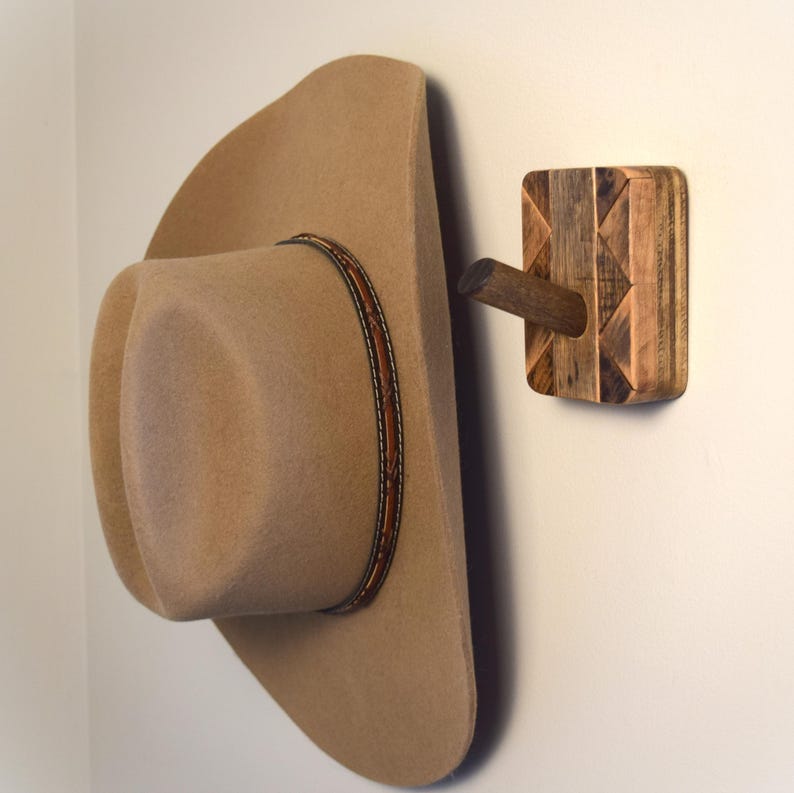 May include: A beige cowboy hat hangs on a wooden wall hook. The hat features a brown leather band with decorative stitching. The hook is constructed from various wood pieces in a geometric pattern, with a wooden peg projecting outwards.