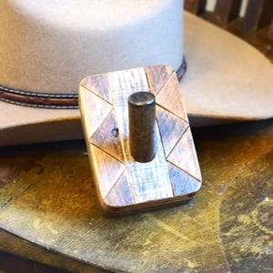 May include: A wooden hat rack with a geometric design. The rack is made of light and dark wood pieces arranged in a triangular pattern. A cylindrical peg extends upwards from the centre. A tan cowboy hat is in the background.