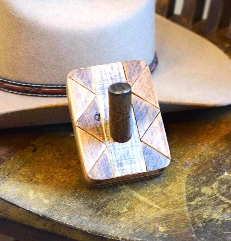 May include: A wooden hat rack with a geometric design. The rack is made of light and dark wood pieces arranged in a triangular pattern. A cylindrical peg extends upwards from the centre. A tan cowboy hat is in the background.