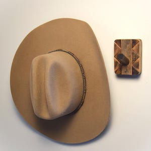 May include: A tan cowboy hat with a decorative band hangs beside a wooden wall hook. The hat has a wide brim and a rounded crown. The hook is made of wood with a geometric design.