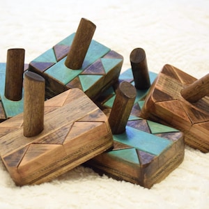 May include: A group of wooden dreidels with geometric patterns. The dreidels have natural wood and teal colour accents. Each dreidel has a cylindrical handle. The items are arranged on a white, textured surface.