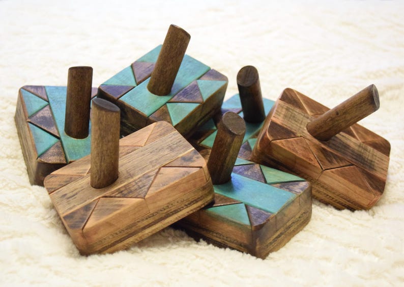 May include: A group of wooden dreidels with geometric patterns. The dreidels have natural wood and teal colour accents. Each dreidel has a cylindrical handle. The items are arranged on a white, textured surface.