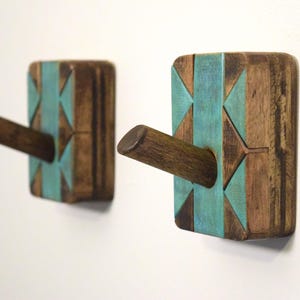 May include: Two wooden wall hooks with a geometric design. The hooks are rectangular with a dark brown finish and teal accents. Each hook has a cylindrical peg extending outward. The hooks are mounted on a white wall.