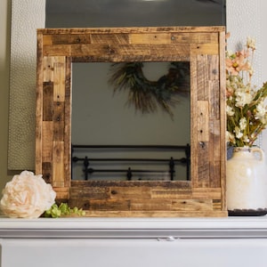 Può includere: Specchio quadrato con cornice in legno di recupero, dal design rustico. La cornice in legno è di varie tonalità di marrone. Un vaso di fiori e un fiore decorativo sono sulla superficie bianca sotto lo specchio.