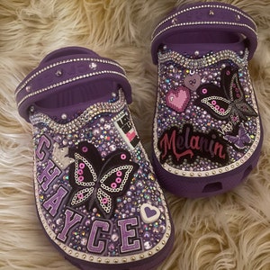 Puede incluir: Un par de Crocs morados adornados con pedrería y adornos. Un zapato presenta el nombre "CHAYCE" y un diseño de mariposa. El otro zapato tiene la palabra "Melanin" y una mariposa. Ambos zapatos están decorados con varias gemas y tachuelas.