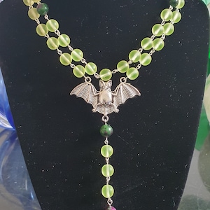 May include: A gothic-style necklace featuring a silver bat pendant and a bird skull charm. The necklace has two strands of light green beads and a strand of green and red beads. The necklace is displayed on a black bust.