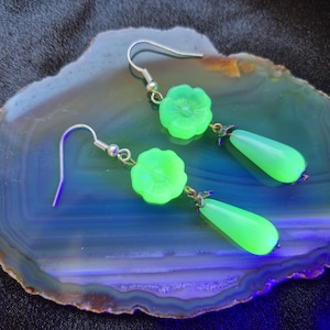 Pode incluir: Um par de brincos verde néon. Cada brinco apresenta um topo em forma de flor e um pendente em forma de gota. Os brincos são suspensos por ganchos de orelha prateados. São exibidos numa pedra azul e castanha.