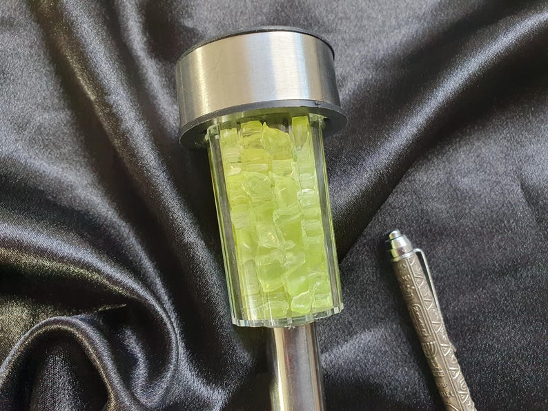 K&ouml;nnte beinhalten: Eine silberne und klare Glassolarleuchte mit einem dekorativen Design aus gr&uuml;nen Glasst&uuml;cken. Die Leuchte befindet sich auf einer schwarzen Stoffoberfl&auml;che.