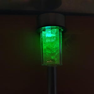 K&ouml;nnte beinhalten: Ein solarbetriebenes Licht aus Silber und Glas mit gr&uuml;nem Leuchten. Das Licht ist so konzipiert, dass es wie ein kleiner Laternenpfahl aussieht.