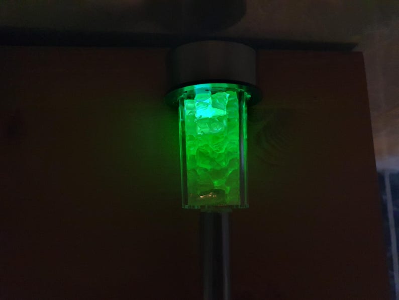 K&ouml;nnte beinhalten: Ein solarbetriebenes Licht aus Silber und Glas mit gr&uuml;nem Leuchten. Das Licht ist so konzipiert, dass es wie ein kleiner Laternenpfahl aussieht.