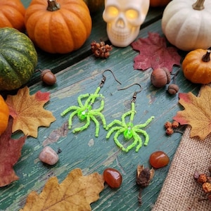 Pode incluir: Um par de brincos pendentes em forma de aranha, feitos com missangas verde néon brilhante e uma conta de cristal verde central para o corpo. Os brincos possuem ganchos de metal escuro para pendurar na orelha.