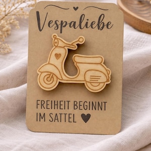Könnte beinhalten: Ein hellbrauner, hölzerner Roller-Ausschnitt auf einer rechteckigen Karte aus Kraftpapier. Die Karte ist mit dem Text "Vespaliebe" oben und "FREIHEIT BEGINNT IM SATTEL" mit einem Herzsymbol unten in schwarzer Schrift bedruckt.