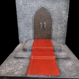 Puede incluir: Una entrada de castillo en miniatura con una alfombra roja que conduce a una puerta de madera. La puerta está situada en una pared de piedra gris con un patrón de ladrillos. La escena es un diorama.