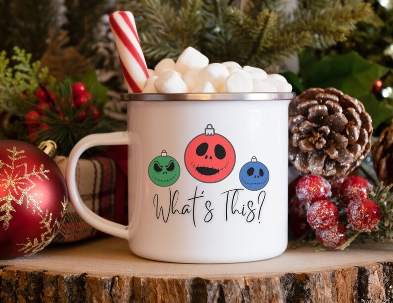 May include: White enamel mug with a red, green, and blue Christmas ornament design featuring the characters from the Nightmare Before Christmas. The text "What's This?" is printed below the ornaments. The mug is filled with marshmallows and a red and white candy cane.