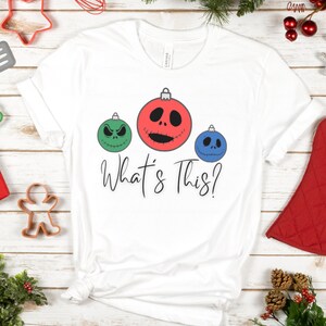 May include: White t-shirt with a green, red, and blue Christmas ornament design. The ornaments have Jack Skellington's face on them. The text "What's This?" is printed below the ornaments.
