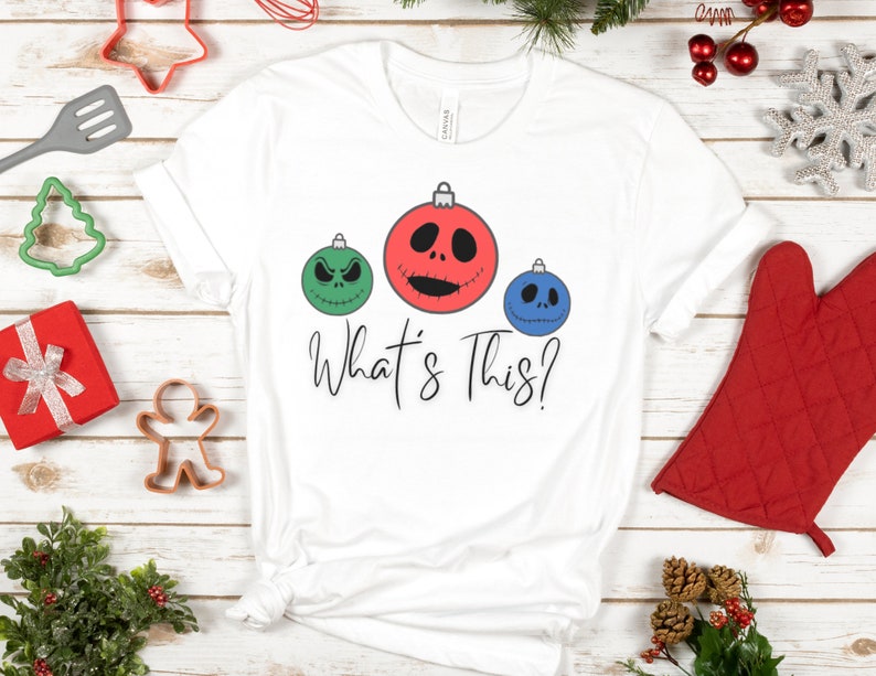 May include: White t-shirt with a green, red, and blue Christmas ornament design. The ornaments have Jack Skellington's face on them. The text "What's This?" is printed below the ornaments.