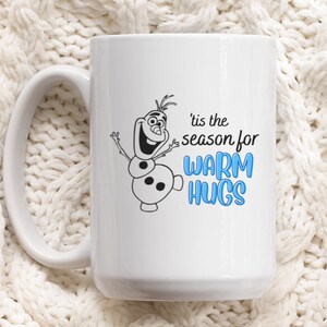 May include: White ceramic mug with a black and white illustration of Olaf from Frozen. The text on the mug reads "'tis the season for warm hugs" in blue.