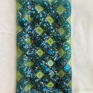 May include: Rectangular glass decorative tray with a textured surface. The tray features a pattern of squares and rounded shapes in shades of teal, blue, and green. The glass has a glossy finish, reflecting light and adding depth to the design.