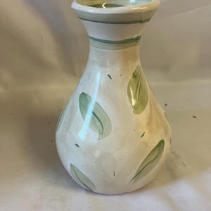 May include: A ceramic vase with a cream-colored base and a hand-painted pattern of light green leaves. The vase features a flared rim with a green horizontal band and a rounded body that tapers toward the neck, showcasing a glossy finish.