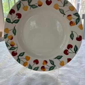 May include: A white ceramic plate with a cherry design. The plate features a border of red and yellow cherries with green leaves. The cherries are arranged around the rim of the plate, creating a decorative pattern.