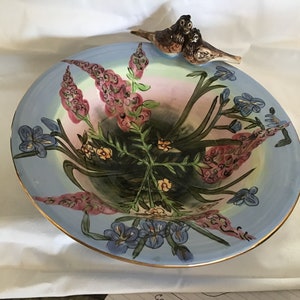May include: A blue ceramic bowl with a gold rim, featuring a hand-painted floral design of pink and blue flowers, green leaves, and two brown birds perched on the rim.