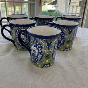 May include: Six ceramic mugs with a blue, green, and white floral pattern. The mugs have a dark blue rim and handle, and a white interior. The mugs are arranged on a white tablecloth.