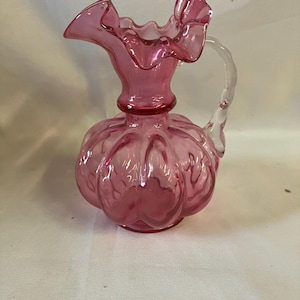 May include: A translucent pink glass pitcher featuring a bulbous, ribbed body and a ruffled, flared rim. The vessel includes a clear, textured glass handle attached to the side, extending from the neck to the shoulder of the rounded base.