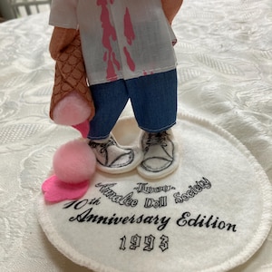 May include: A doll figurine with a white shirt featuring pink splatters, blue jeans, and white shoes. The doll holds a brown ice cream cone with pink ice cream. The base reads "Tinakee Doll Society 10th Anniversary Edition 1993."