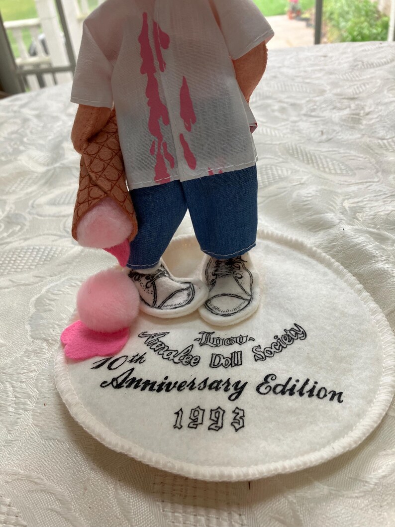 May include: A doll figurine with a white shirt featuring pink splatters, blue jeans, and white shoes. The doll holds a brown ice cream cone with pink ice cream. The base reads "Tinakee Doll Society 10th Anniversary Edition 1993."