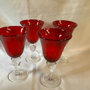 May include: A set of four matching goblets featuring deep red glass bowls and clear glass stems with a twisted design. Each goblet has a wide, flared rim and a circular base, showcasing a consistent color and structural style across all pieces.