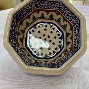 May include: An octagonal ceramic bowl with a cream-colored exterior and a detailed interior design. The interior features a blue star pattern, green trees, and brown and blue decorative borders. The bowl is likely for serving or display.