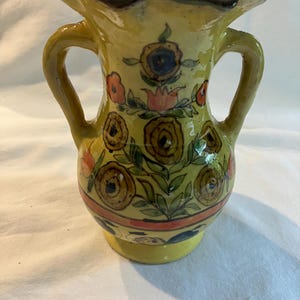 May include: A hand-painted ceramic vase with two handles, featuring a yellow base and body. The vase is adorned with floral designs in shades of brown, orange, and green. The rim is green with a brown trim.