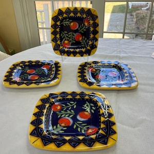 May include: Four square ceramic plates with a fruit design. The plates have a navy blue center with red and white fruit and green leaves. The edges are yellow with a geometric pattern.